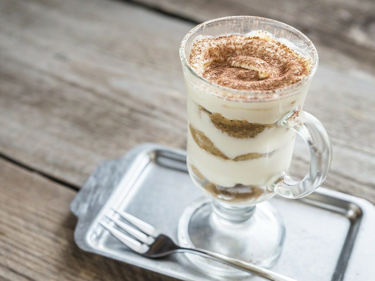 Eine Portion Bananen-Tiramisu un einem Glas steht auf einem silbernen Tablett auf einem braunen Holztisch. Neben dem Glas liegt eine silberne Gabel. Das Tiramisu ist angerichtet mit Kakaopulver.