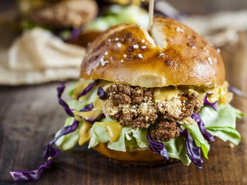 Ein Burger mit Blumenkohl-Patty steht auf einem raunen Holzbrett auf einem braunen Untergrund. Im Hintergrund steht ein weiterer Burger.