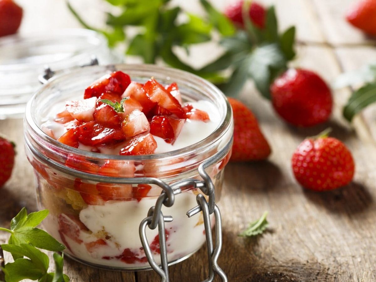 Eine Portion Erdbeer-Tiramisu im Glas steht auf einem Holztisch. Das Tiramisu ist angerichtet mit Erdbeerstückchen. Im Hintergrund liegen einzelne Erdbeeren und Grün.