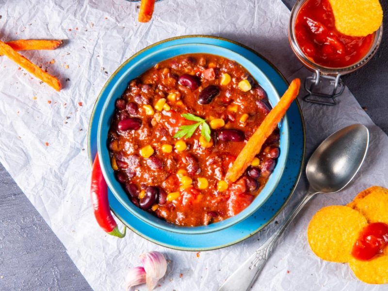 Auf einem rustikalem Tisch liegt ein helles Tuch. Darauf steht eine große Schüssel mit blauem Rand, in der das Süßkartoffel-Chili zu sehen ist. Neben der Schüssel liegen noch Süßkartoffel-Sticks, sowie einige Nachos und ein Glas mit einer roten Soße.