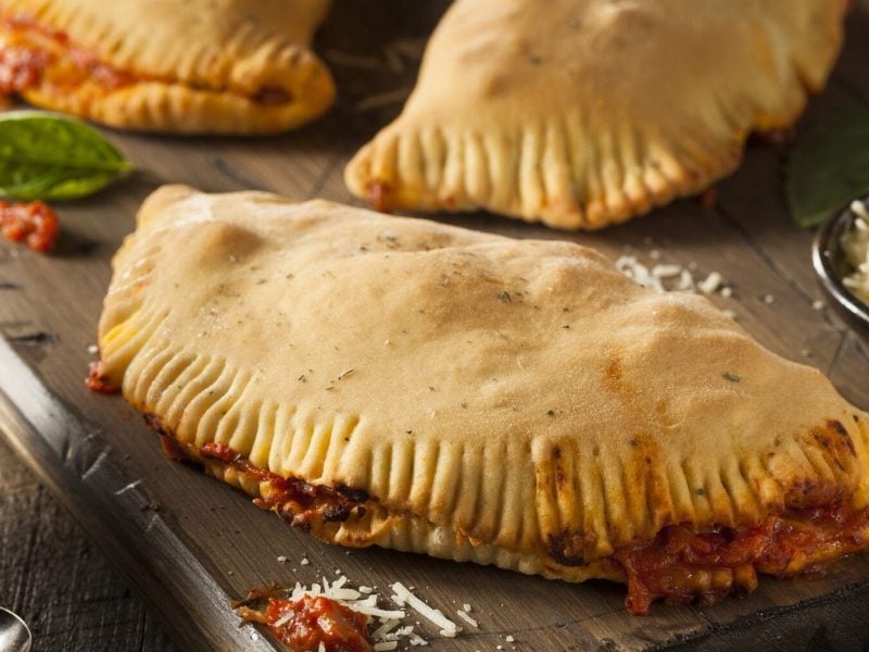 3 Gemüse-Schinken-Calzones liegen auf einem Holzbrett. Links neben dem Brett steht ein Gefäß mit Tomatensoße, rechts eine kleine Schale mit Käse.