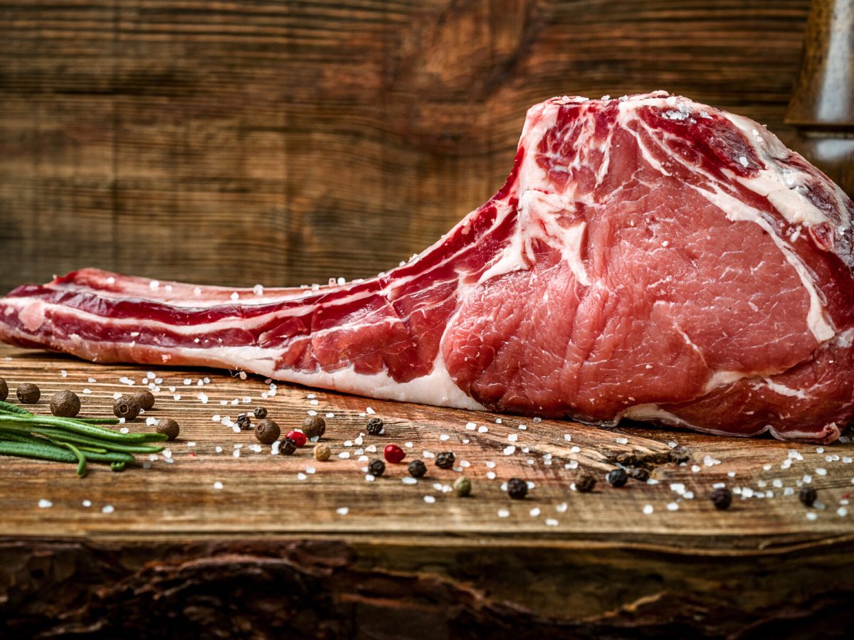 Ein saftig marmoriertes Tomahawk-Steak liegt mit Meersalz und Pfefferkörnern auf einem Holzuntergrund.