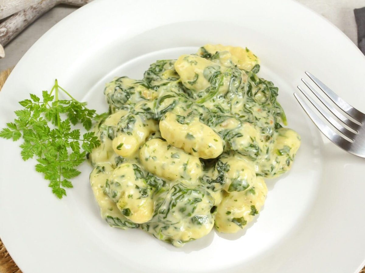 Eine Portion Gnocchi mit Spinat-Béchamel auf einem weißen Teller steht auf einer geflochtenen Matte auf einem Tisch. Am rechten Rand vom Teller liegt eine silberne Gabel.