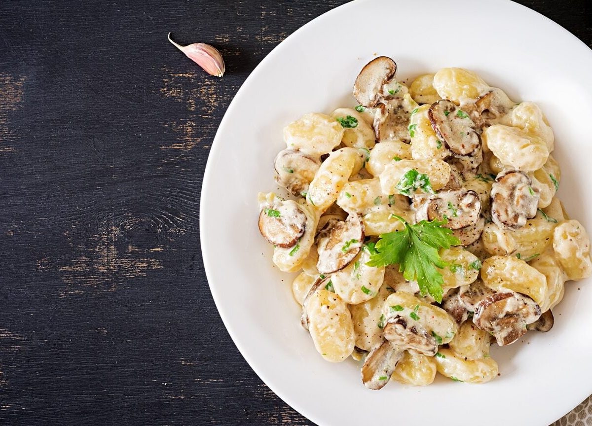 Ein weißer Teller mit Gnocchi con Funghi steht auf einem braunen Holztisch. Neben dem Teller liegt ein gepunktetes Geschirrtuch und eine Knoblauchzehe.