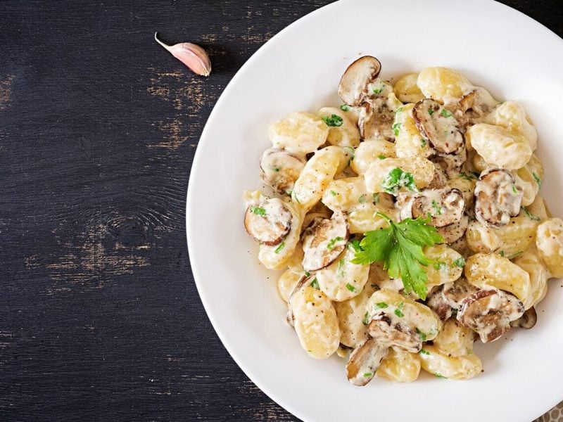 Ein weißer Teller mit Gnocchi con Funghi steht auf einem braunen Holztisch. Neben dem Teller liegt ein gepunktetes Geschirrtuch und eine Knoblauchzehe.