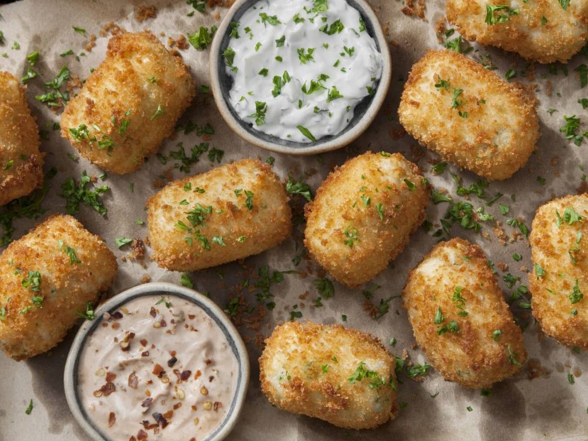 Mehrere Kroketten mit Petersilie liegen auf einem Blech mit Backpapier. Neben den Kroketten stehen verschiedene Dips.