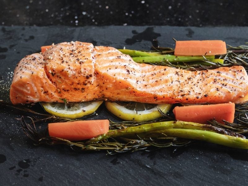 Ein Stück Lachsfilet liegt auf Rosmarin, Zitronenscheiben, Spargel und Karotten. Der Untergrund ist eine schwarze Steinplatte. Über dem Lachs und dem Gemüse sind Salz und Pfefferkörner verteilt.