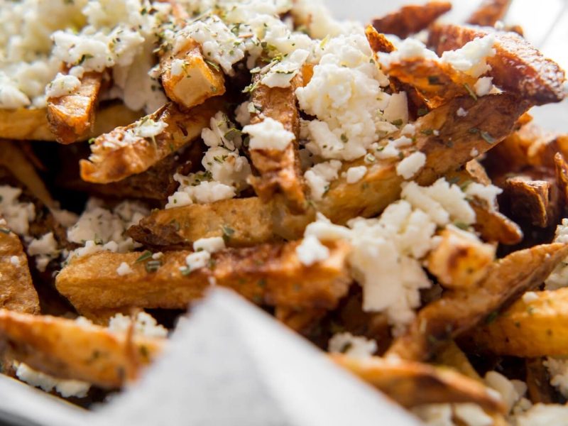 Eine Portion Loaded Fries mit Feta liegt in einer mit einer Serviette ausgelegten Metallschüssel. Die Loaded Fries sind mit verschiedenen Gewürzen bestreut.