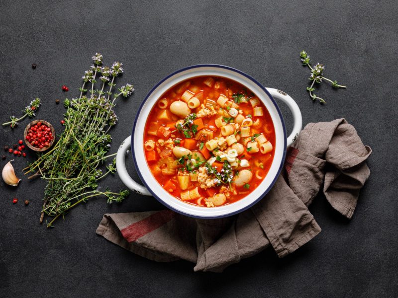 Italienische Minestrone mit Pasta und Bohnen angerichtet auf dunkler Schieferplatte, umgeben von Zutaten. Platte