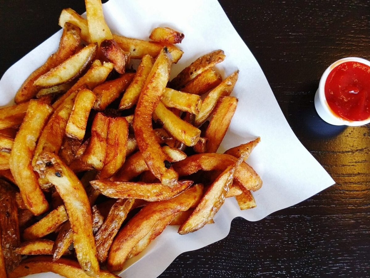 Eine Portion Pommes auf einer weißen Servierte liegt auf einem schwarzen Holztisch, Neben den Pommes steht ein kleiner Becher mit Ketchup.