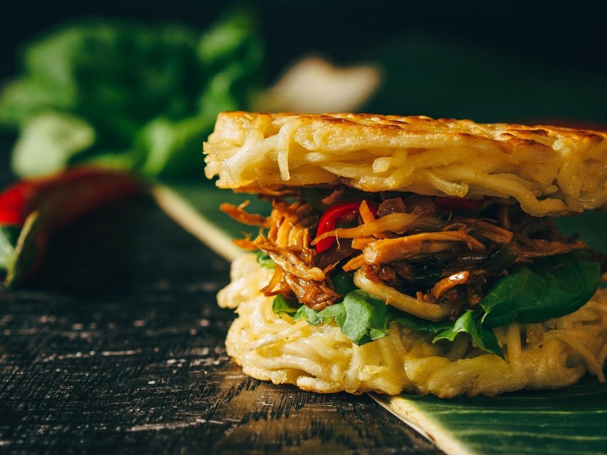 Ein Ramen-Burger steht auf einem Bananenblatt auf einem schwarzen Tisch. Der Ramen-Burger ist belegt mit Tomaten, Rucola, Hähnchen und Zwiebeln. Im Hintergrund liegt Salat und mehrere Chilichoten.