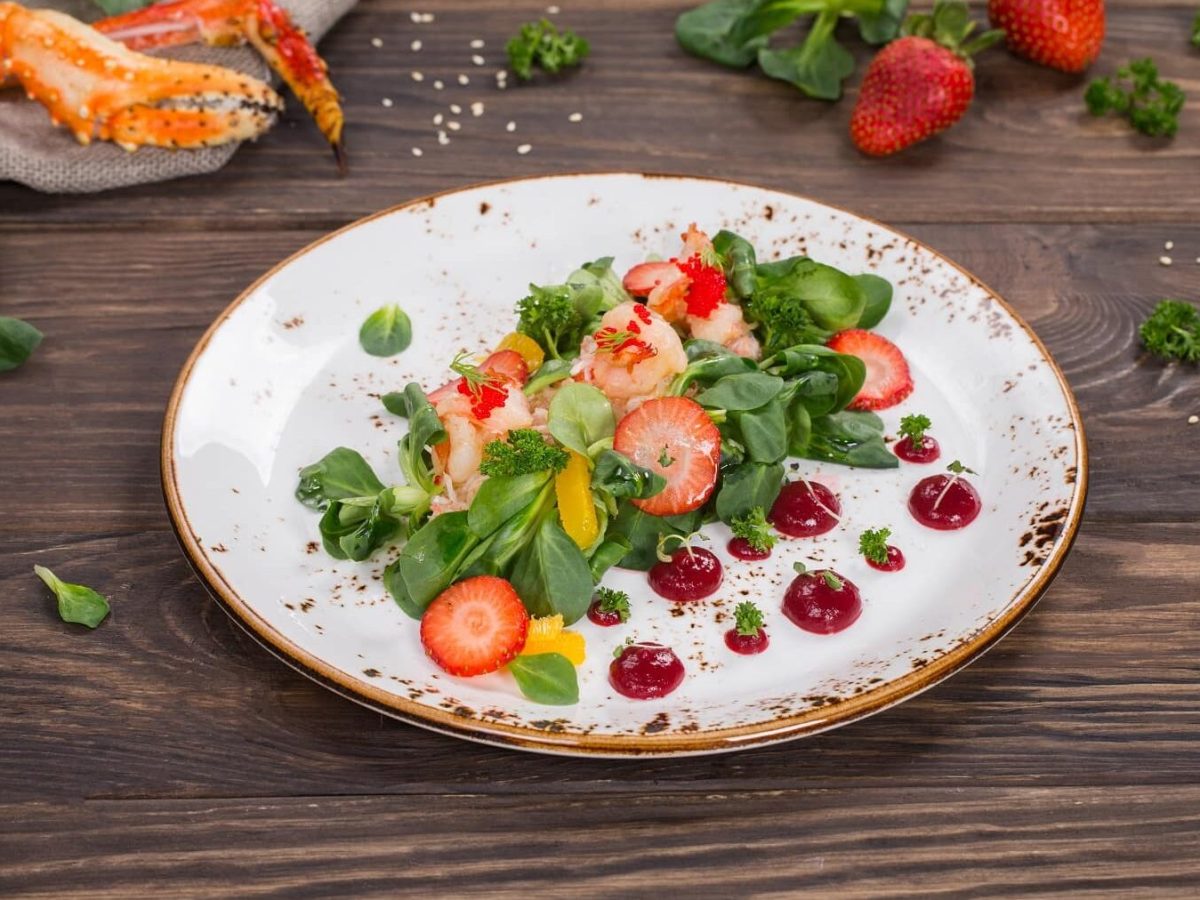 Ein weißer Teller mit Shrimps-Salatmit Erdbeeren steht auf einem braunen Tisch. Auf dem Tisch liegen noch Krabben, Salat, Petersilie und Erdbeeren.