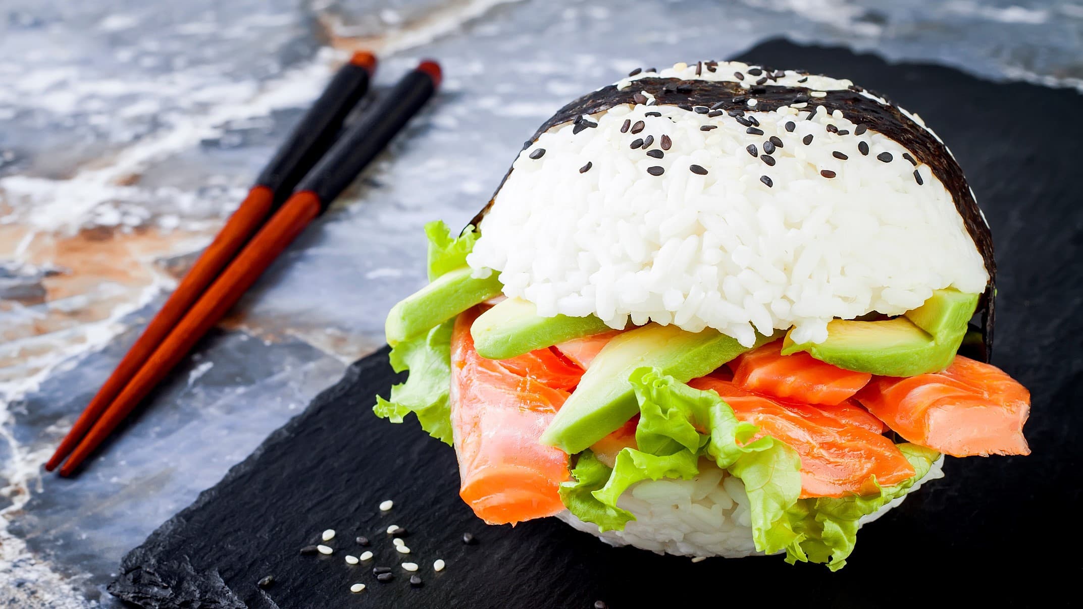 Ein Sushi-Burger mit Avocado und Lachs auf einer schwarzen Schiefernplatte. Daneben liegen Stäbchen.