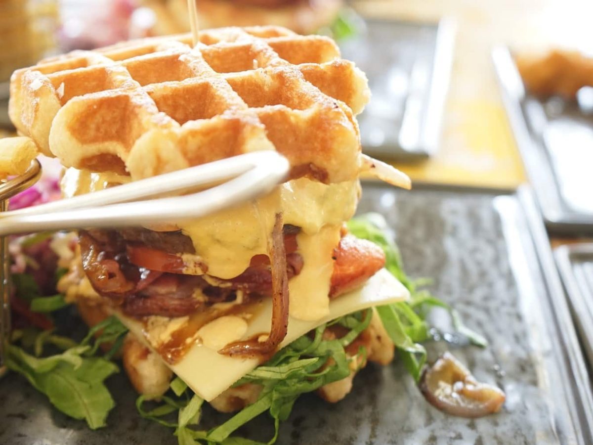 Ein Waffel-Burger liegt auf einer Steinplatte. Der Burger ist belegt mit Zwiebeln, Tomaten, Rucola, Tomaten, einem Burgerpatty und viel Käse. Neben dem Burger steht eine Portion Pommes.