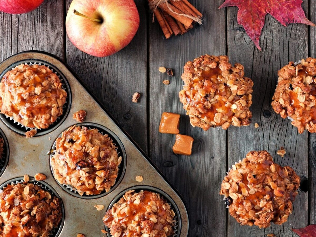 Drei Apfel-Karamell-Streusel-Muffins liegen auf einem grauen Holztisch. Neben ihnen steht eine Muffinform mit weiteren Muffins. Auf dem Tisch verteilt liegen Caramelwürfel, Haferflocken, Äpfel und Zimtstangen.