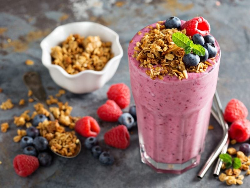 Ein Glas Aronia-Smoothie, dekoriert mit Müsli und frischen Beeren. Drumherum liegen verstreut die Zutaten.