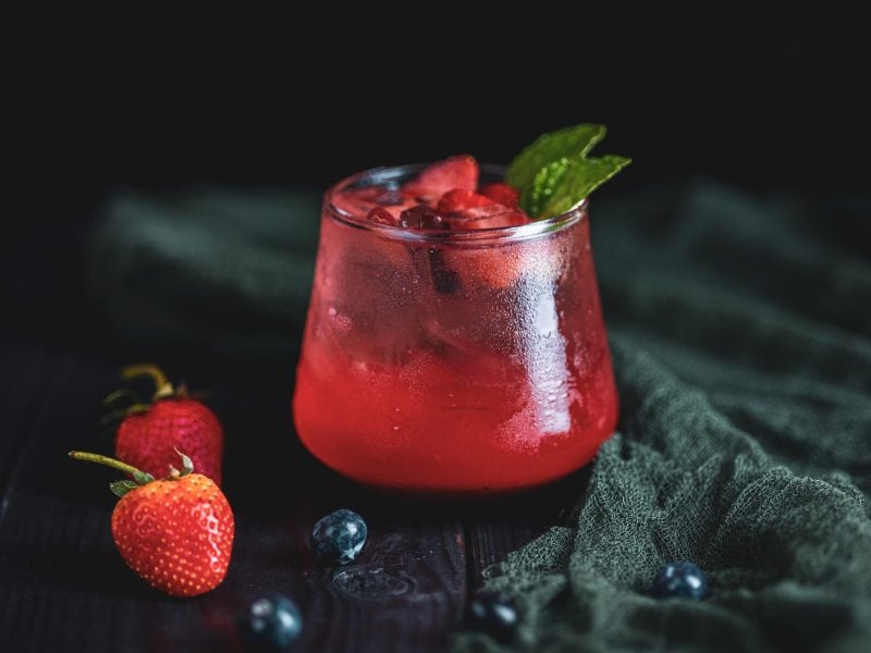 Ein Glas mit rotem Berlin Berry Cocktail, dekoriert mit frischen Beeren, vor einem dunklen Hintergrund.