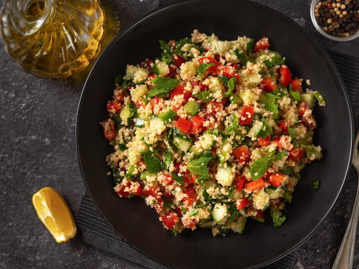 Ein dunklter Teller mit einer Portion Couscous-Tomatensalat, daneben Besteck, eine Zitronenspalte, eine Flasche mit Öl und eine Schale mit Pfefferkörnern.