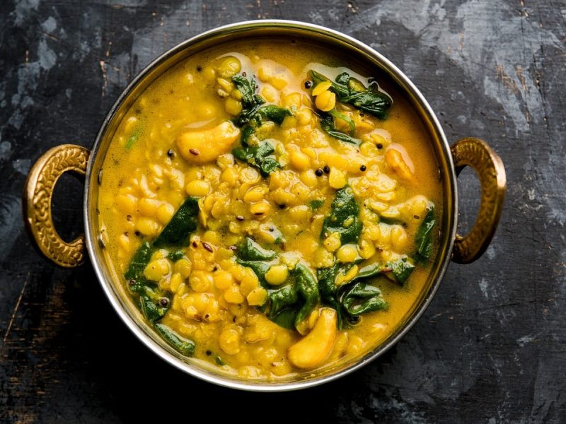 Eine gusseiserne Schüssel mit verzierten Henckeln gefüllt mit gelben Dal Palak, Linsen-Curry mit Spinat, hier verfeinert mit Cashew-Nüssen und Kreuzkümmel, vor einem dunklen Hintergrund von oben.