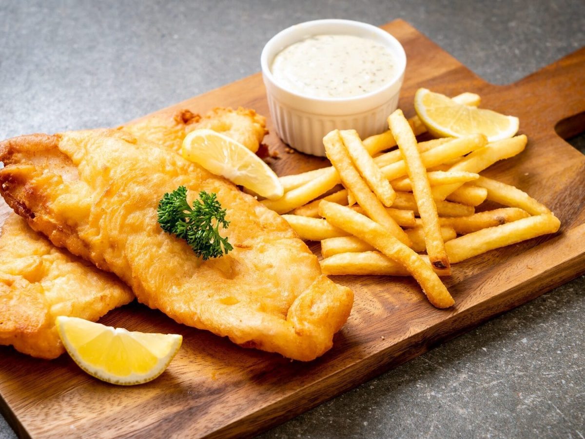 Eine Portion Fish and Chips mit Dip auf Holzbrett, angerichtet auf dunklem Untergrund