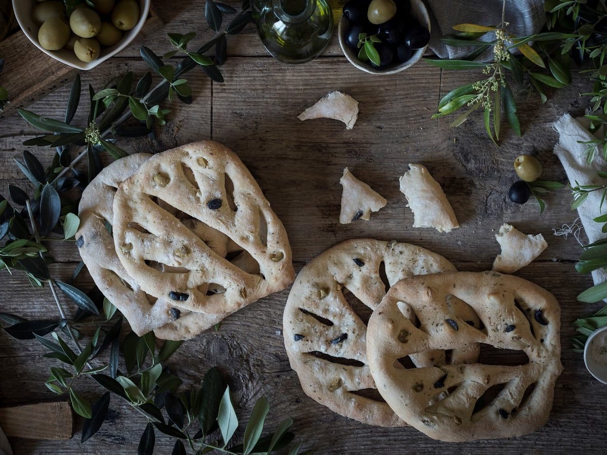 Vier Laib des französischen Brotes Fougasse, deren Form an Blätter erinnert, nebeneinander und aufeinander gestapelt, umrandet von Blättern, Tüchern und Oliven, vor einem dunklen Holzhintergrund, von oben.