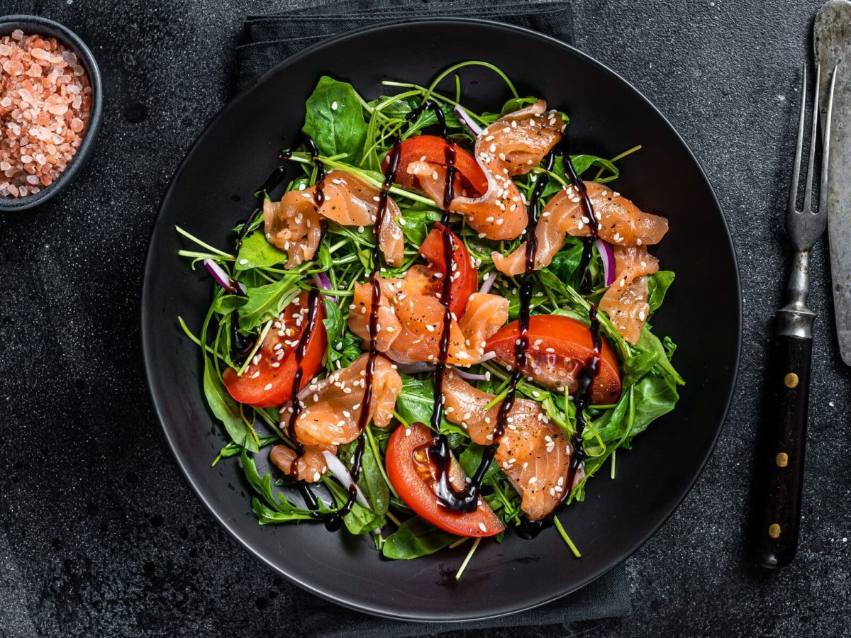 Ein dunkler Teller mit grünem Salat mit Lachs und Tomaten. Daneben liegt Besteck, auf der anderen Seite steht eine Schale mit grobem Salz.