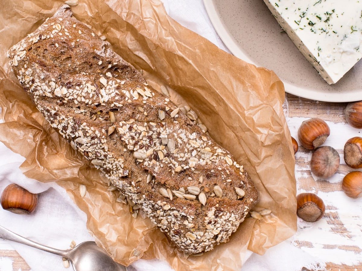 Ein Laib Haselnussbrot mit Haferflocken und Kürbiskernen bestreut auf einem Stück Backpapier neben Haselnüssen und Käse von oben vor einem hellen Holz Hintergrund.