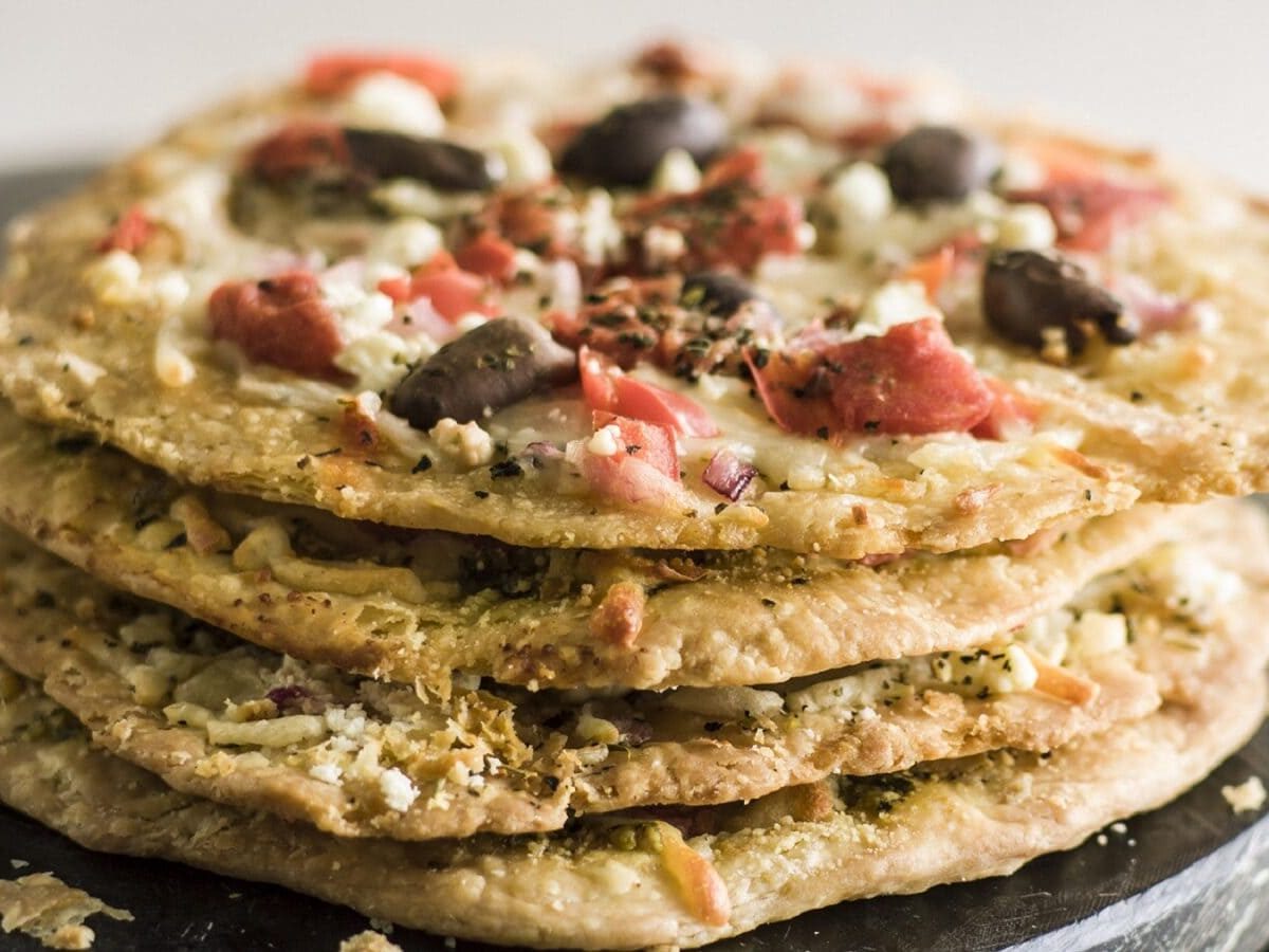 Vier Hüttenkaese-Taler mit Oliven und Tomaten liegen uebereinander auf einem schwarzen Brett auf einem grauen Untergrund.