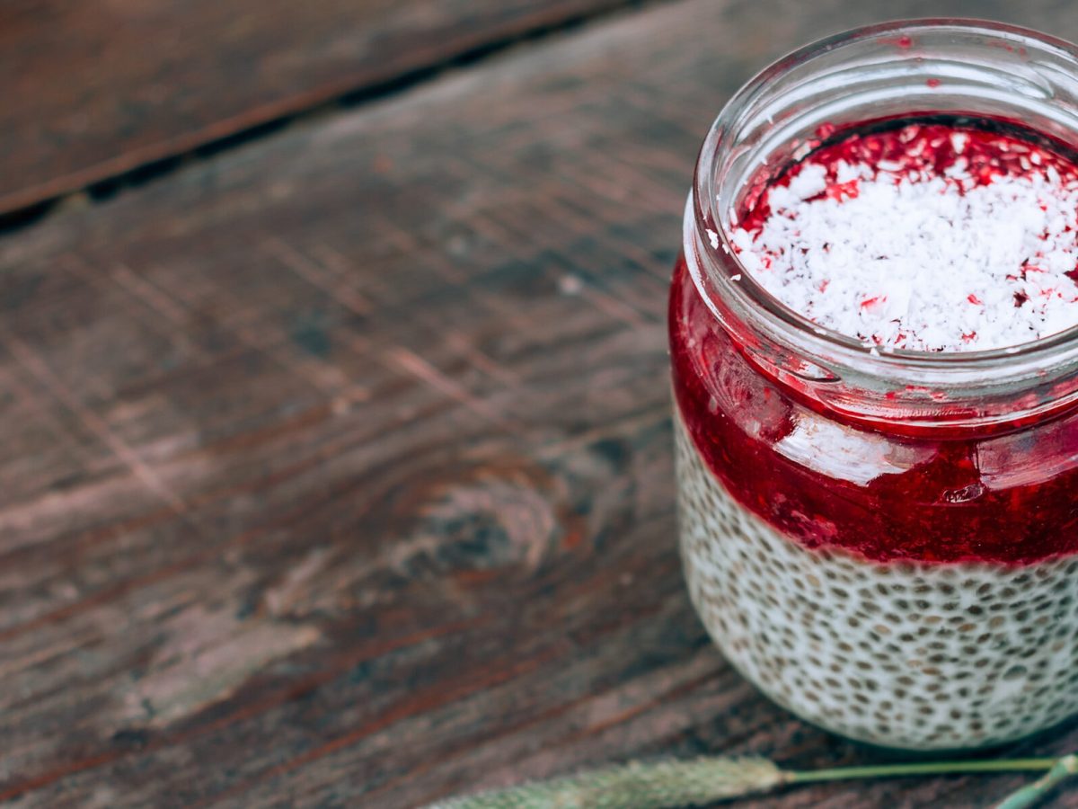 Ein Glas mit Kokos-Chia-Pudding, getoppt mit Himbeerpüree und Kokosraspeln, auf einem Holztisch.