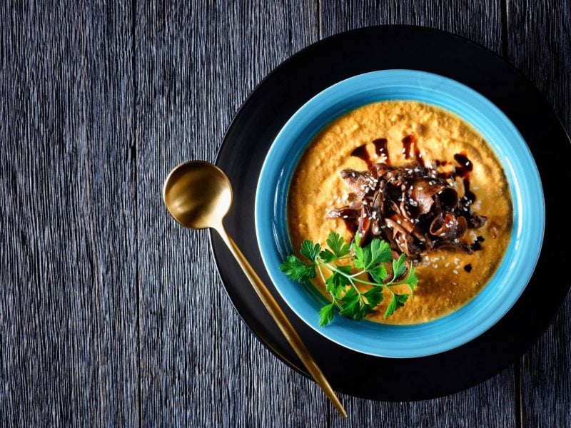 Ein blauer Teller mit Kürbissuppe mit Pilzen und Balsamico sowie Sesamkörnern, daneben ein goldener Löffel. Alles auf einem Holztisch in der Draufsicht.