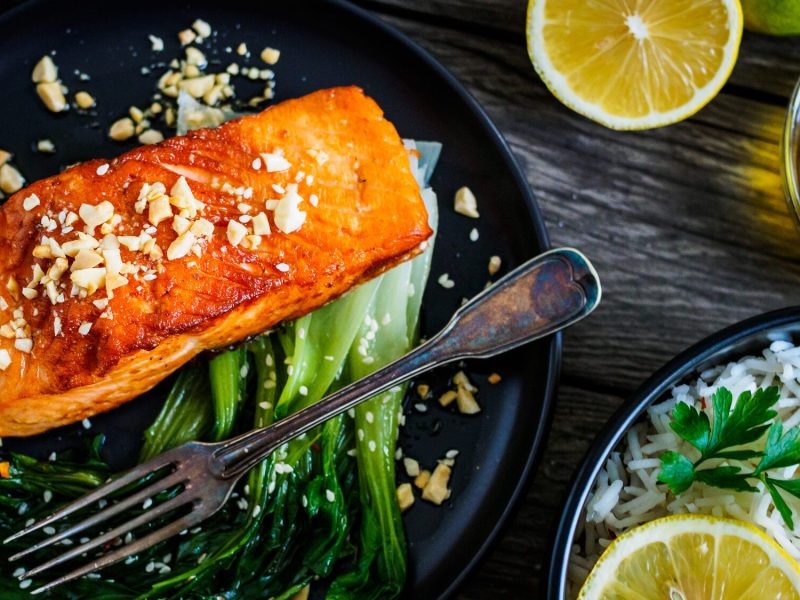 auf einem Holztisch steht ein dunkler Teller. Auf dem Teller liegt ein Lachsfilet mit Pak Choi. Auf dem Tisch liegen Zitronenscheiben.