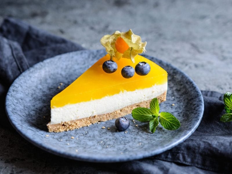 Ein Teller mit einem Stück Mango-Cheesecake, dekoriert mit Blaubeeren und Physalis.