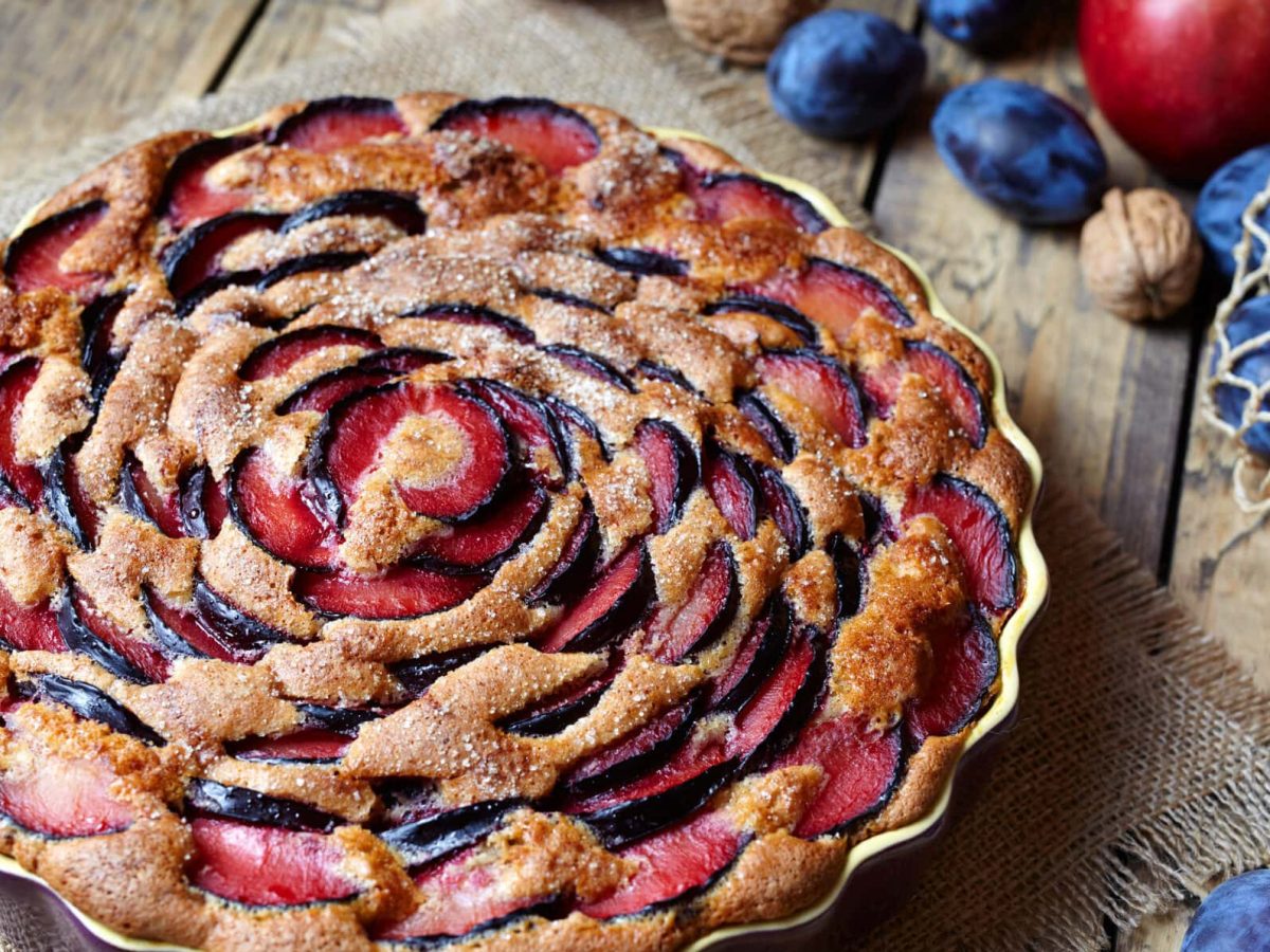 Eine Kuchenform mit Pflaumenkuchen mit Joghurt, daneben liegen Zwetschgen, Äpfel und Nüsse.