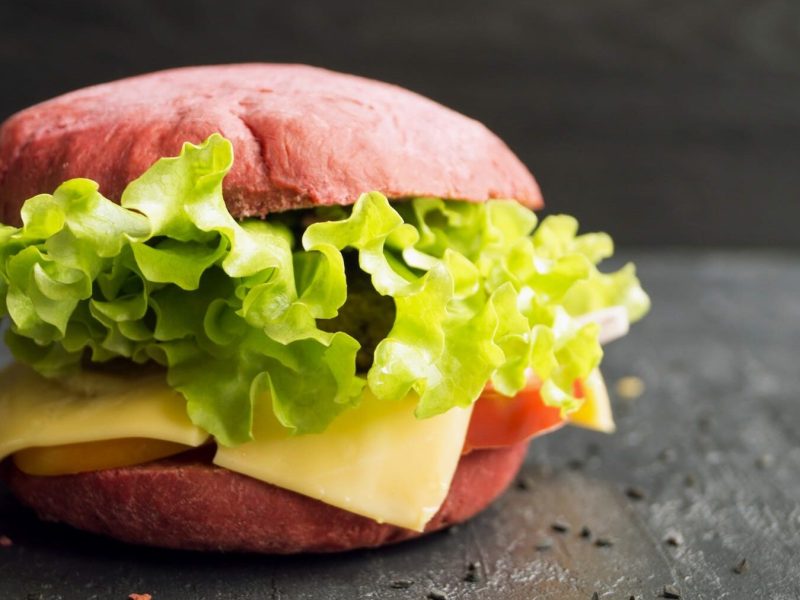 Ein Burger mit pinken Burger Buns, mit Salat, Käse und Tomaten steht auf einem schwarzen Holztisch. Auf dem Tisch verteilt liegen Sesamsamen. Der Hintergrund ist ebenfalls schwarz.