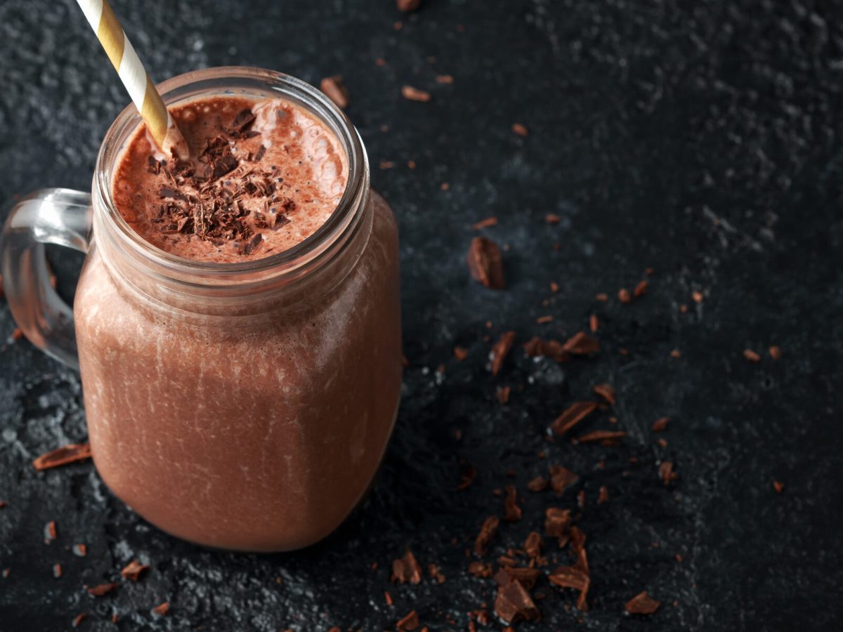 ein Glas Proteinshake mit Schokolade und einem Strohhalb vor dunklem Hintergrund.