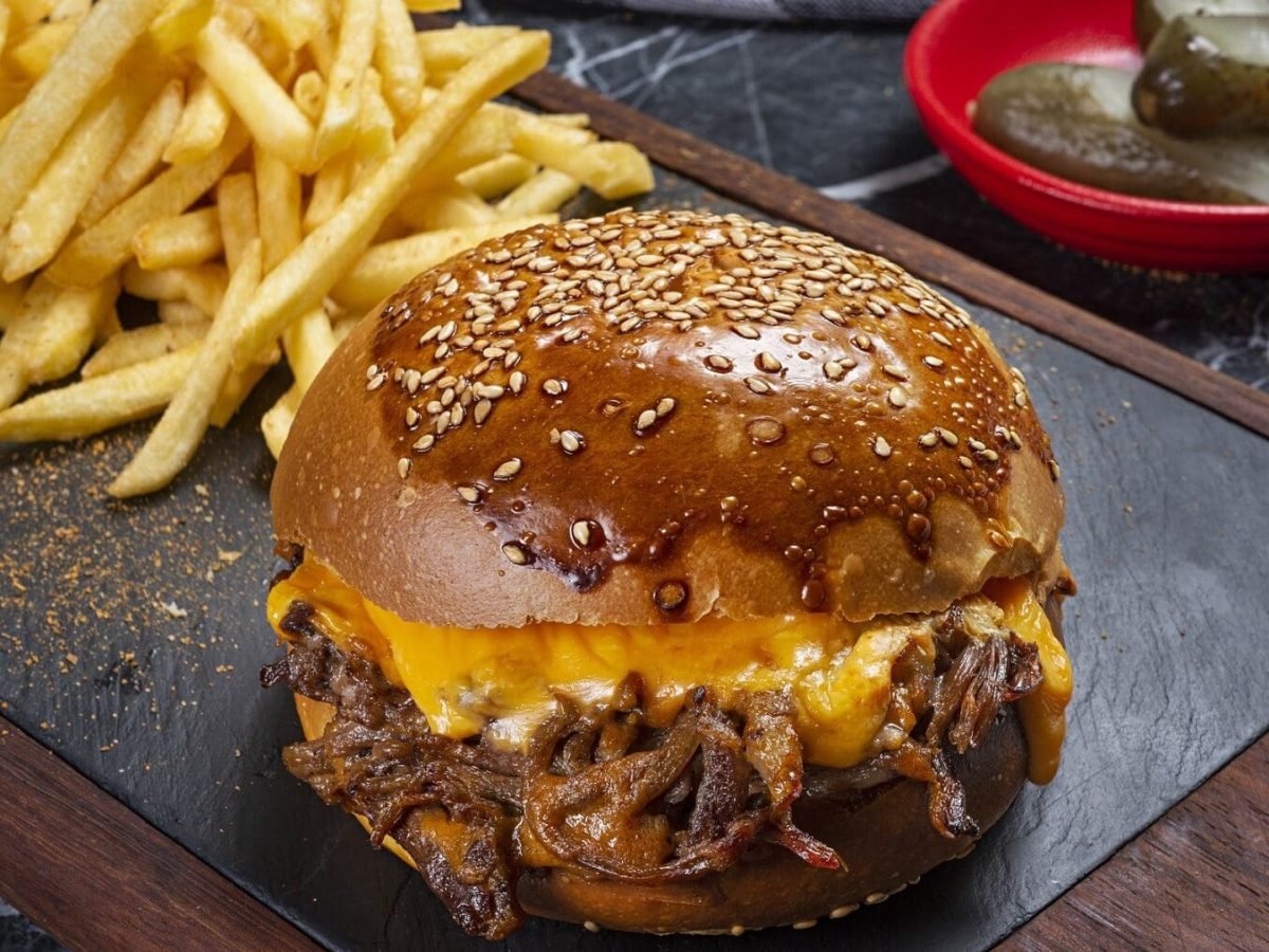 Ein Pulled Beef Burger mit Käsesauce steht auf einer Steinplatte auf einem schwarzen Tisch. Neben dem Burger liegen Pommes. Im Hintergrund steht eine rote Schale mit Gewürzgurken.