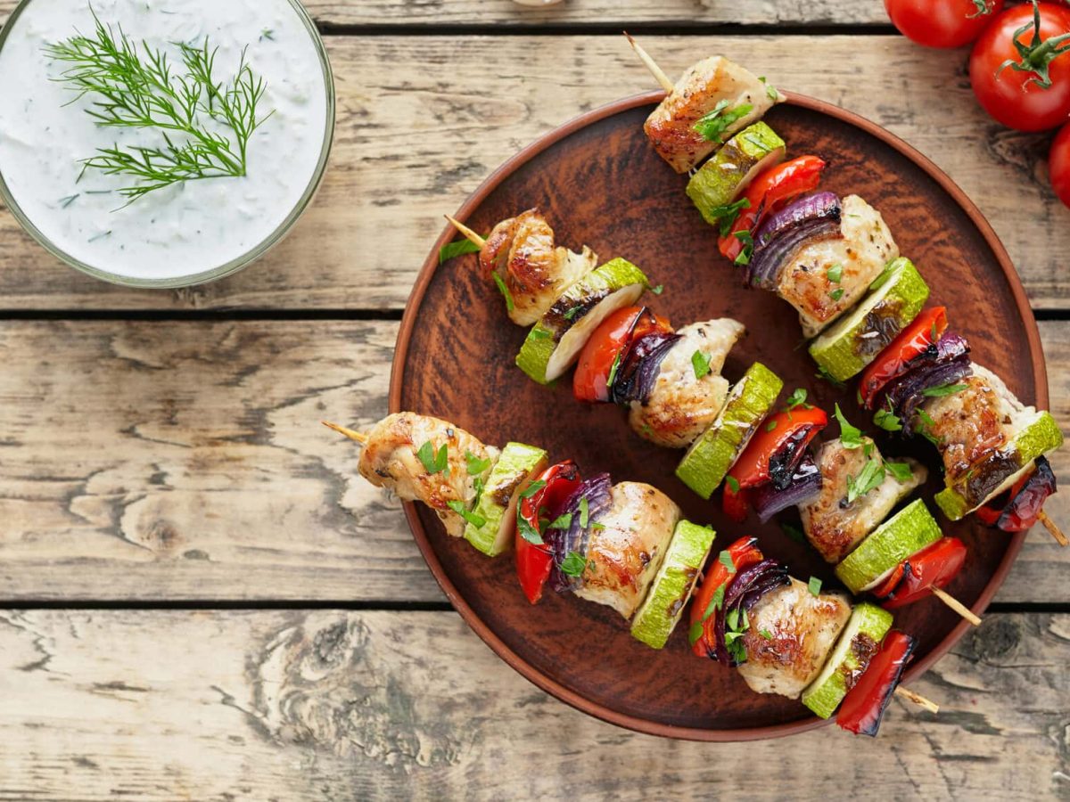 Putenspieße mit Gemüse auf einem dunklen Teller in der Draufsicht. Daneben liegen Strauchtomaten und Knoblauch, außerdem ist eine Schale mit Quark zu sehen.