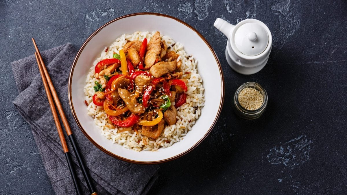 Ein Teller Reis mit Hähnchen und Paprika, daneben liegen Essstäbchen und ein Geschirrtuch. Auf der anderen Seite stehen ein Kännchen sowie ein Glas mit Sesam.