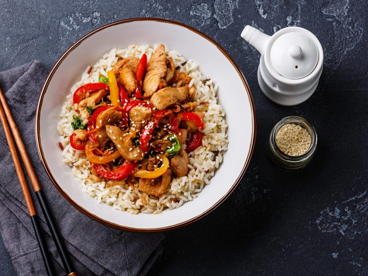 Ein Teller Reis mit Hähnchen und Paprika, daneben liegen Essstäbchen und ein Geschirrtuch. Auf der anderen Seite stehen ein Kännchen sowie ein Glas mit Sesam.