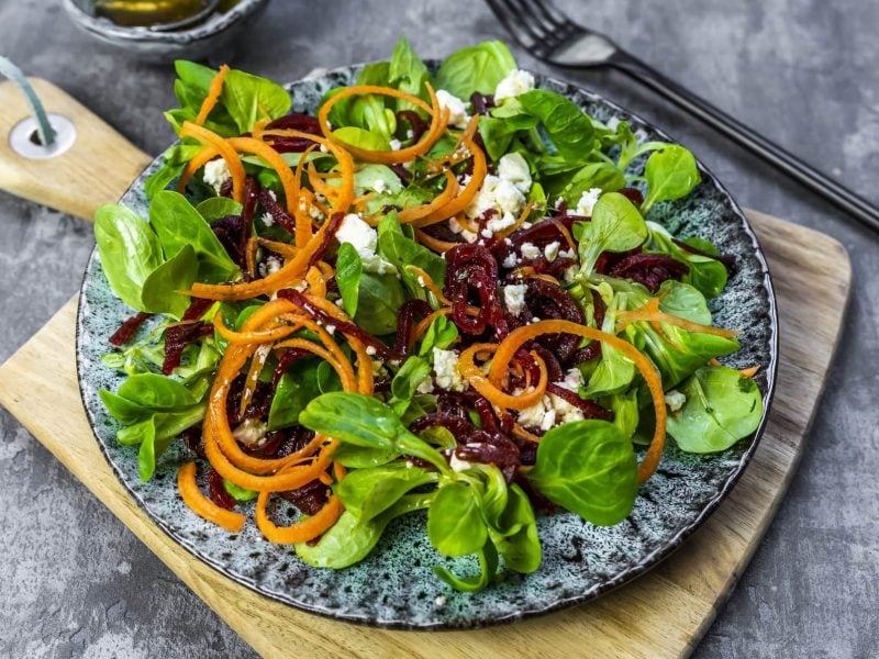 Rote-Bete-Salat mit Feldsalat und Feta auf einem gemusterten Teller. Der Teller steht auf einem Holzbrett. Im Hintergrund liegt eine schwarze Gabel.