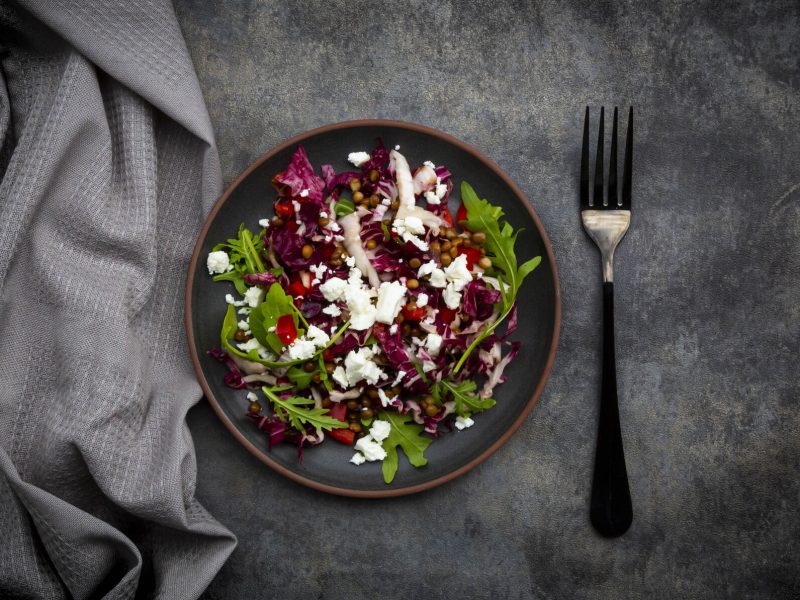 Ein Teller mit Rucola-Radicchio-Salat, Feta und Linsen, daneben liegen eine Gabel und eine graue Stoffserviette.