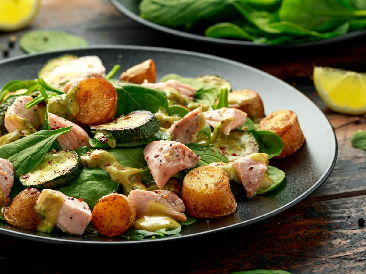 Ein Teller Salat mit Lachs, Blattspinat und Kartoffeln, daneben liegen Spinatblätter und Zitronenscheiben.