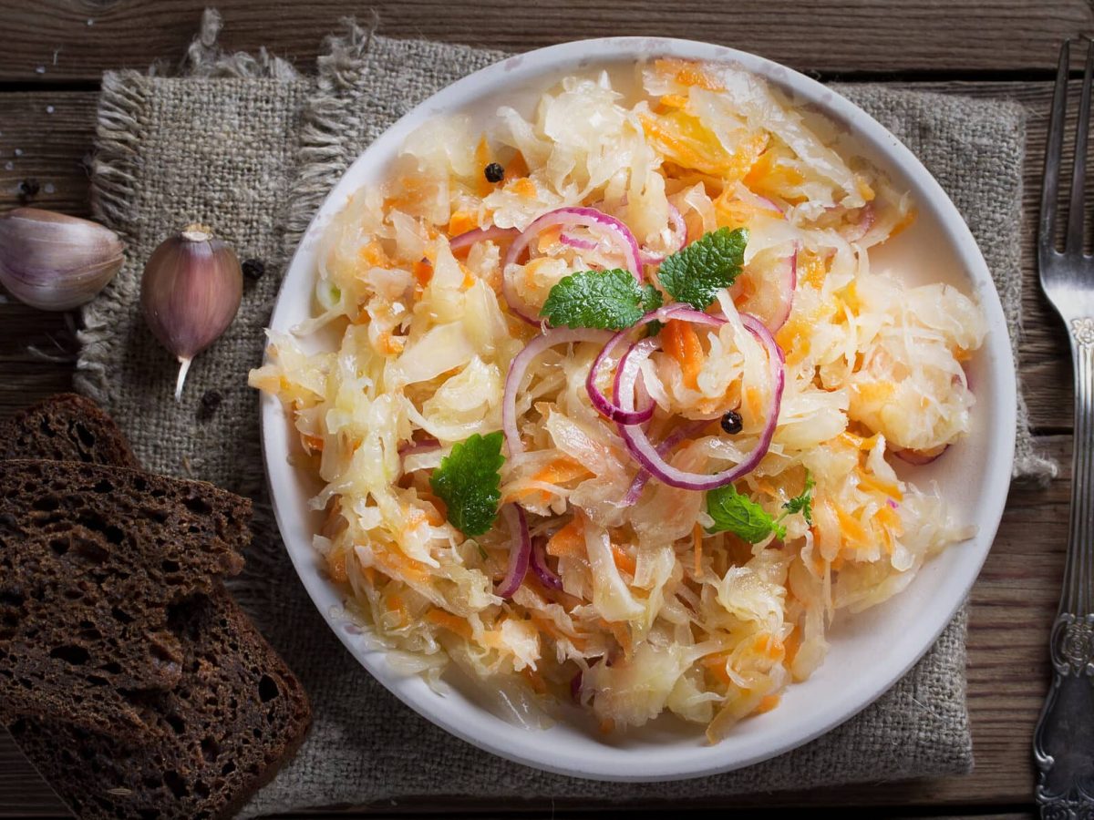 ein heller Teller mit Sauerkrautsalat, daneben liegt eine Gabel, auf der anderen Seite sieht man Knoblauchzehen, Salz und Schwarzbrot. Alles auf Holz.