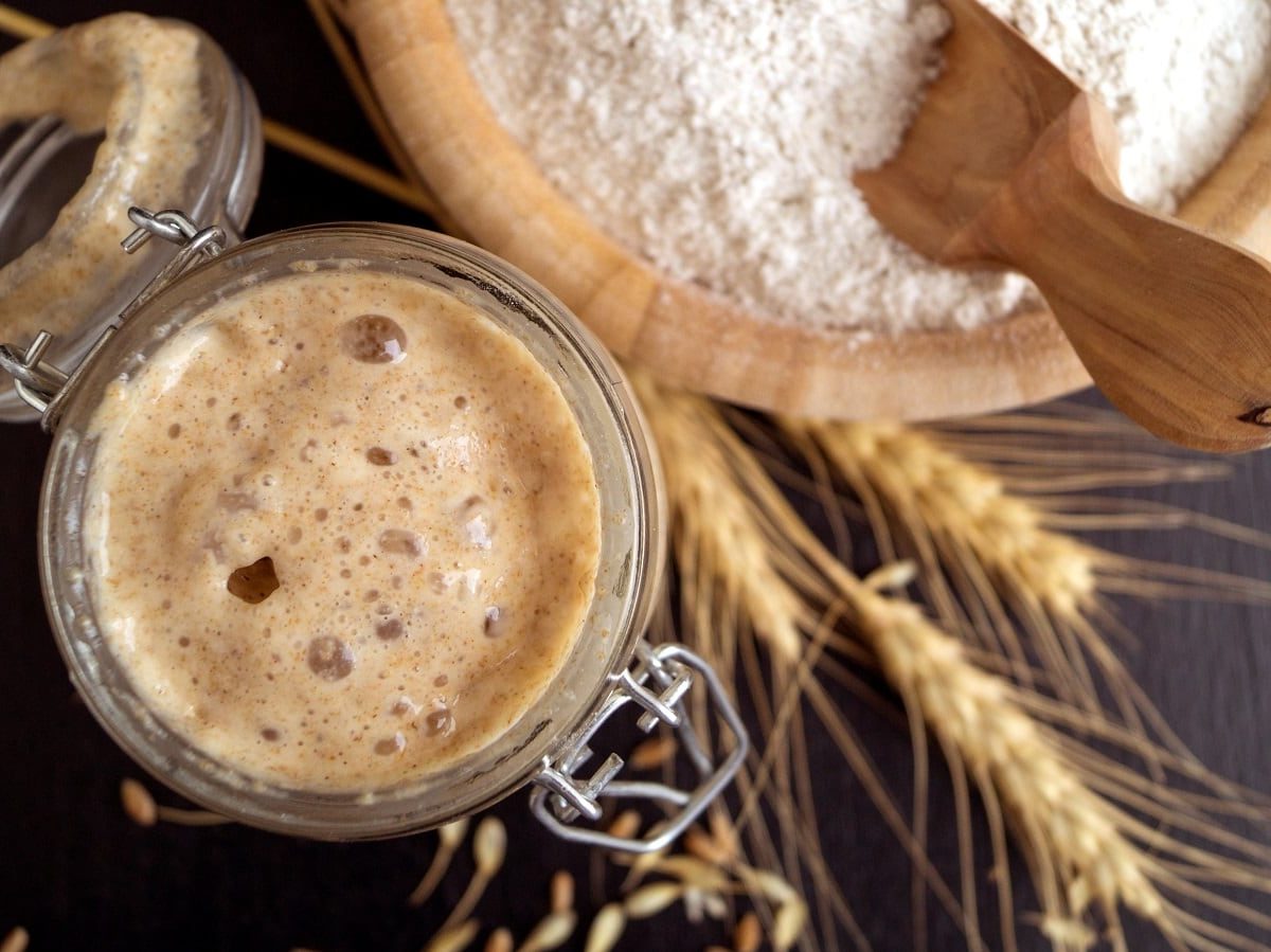 Angesetzter Sauerteig in einem geöffneten Glas neben einer Holzschüssel mit Mehl und Weizen-Ähren, von oben, vor dunklem Hintergrund.