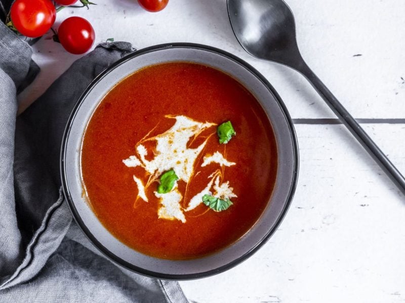 eine Schüssel mit schneller Tomatensuppe, verfeinert mit Basilikum und Joghurt. Daneben liegen Tomaten, ein Geschirrtuch und ein Löffel.