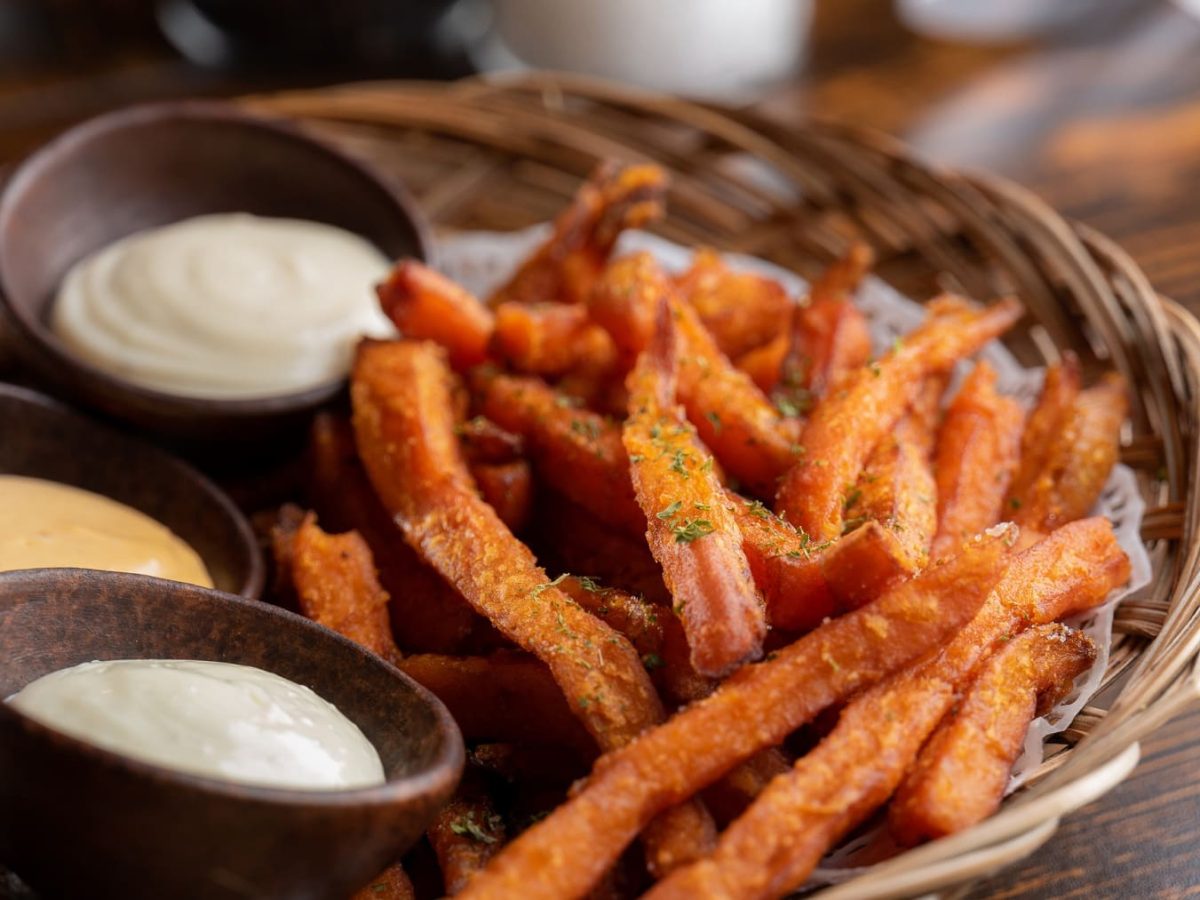 Eine Portion Süßkartoffel-Pommes liegt in einem kleinen Körbchen. Neben den Pommes stehen drei braune Schalen mit verschiedenen Dips. Das Körbchen steht auf einem braunen Holztisch.