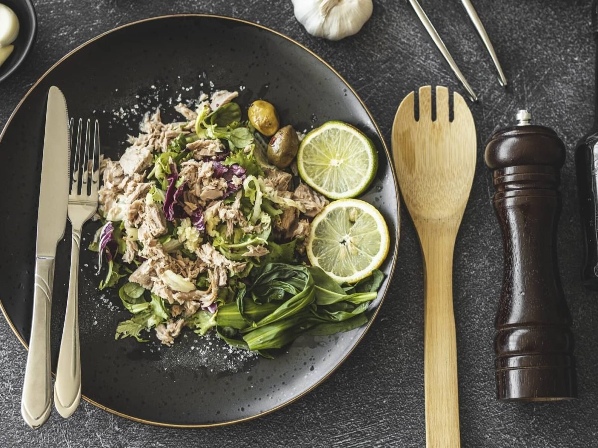 Ein dunkler Teller mit Thunfischsalat mit Oliven, daneben liegt Besteck, außerdem eine Gewürzmühle, eine Ölflasche und ein paar Zutaten.