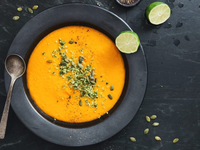 Ein dunkler Teller auf einem dunklen Tisch in der Draufsicht, darin vegane Kürbissuppe, mit Kernen bestreut. Daneben liegen zwei Limettenhälften und ein Löffel.