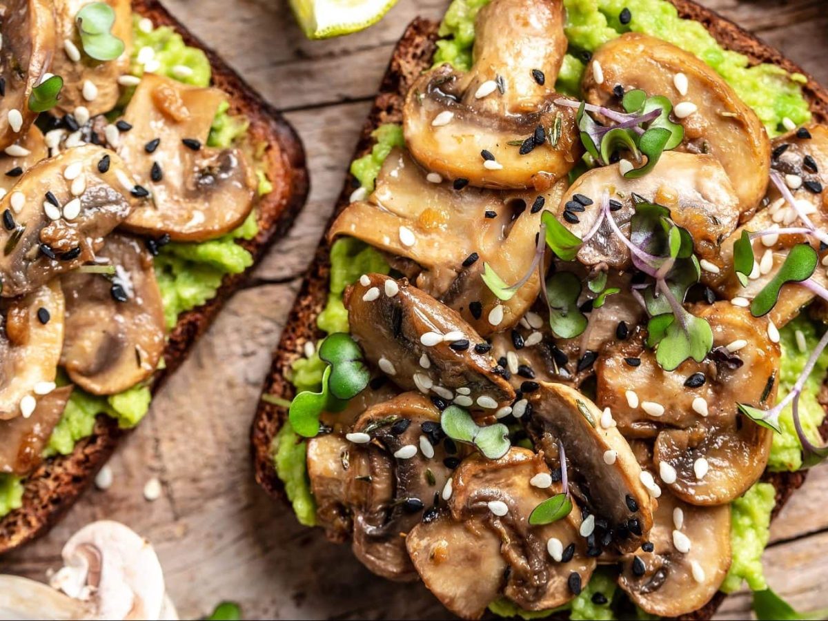 Zwei Scheiben Avocado-Toast mit Pilzen auf einem braunen Holztisch. Um die Brote herum liegen Limettenscheiben und Pilze. Das Avocado-Toast ist angerichtet mit Sesam und Sprossen.