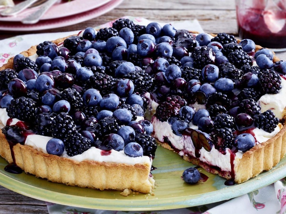 Eine Brombeer-Blaubeer-Tarte auf einem bunten Teller auf einem Holztisch. Um den Teller herum steht ein Glas mit Brombeer-Gelee, Geschirr und Servietten mit Blumendruck.